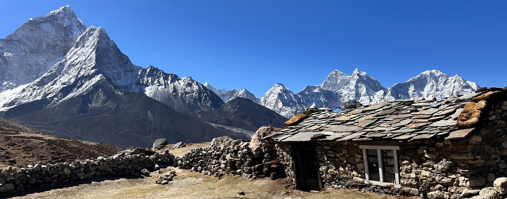 Traditional stone Sherpa hut with a slate roof beneath towering snow-capped Himalayan mountains on the EBC trail.